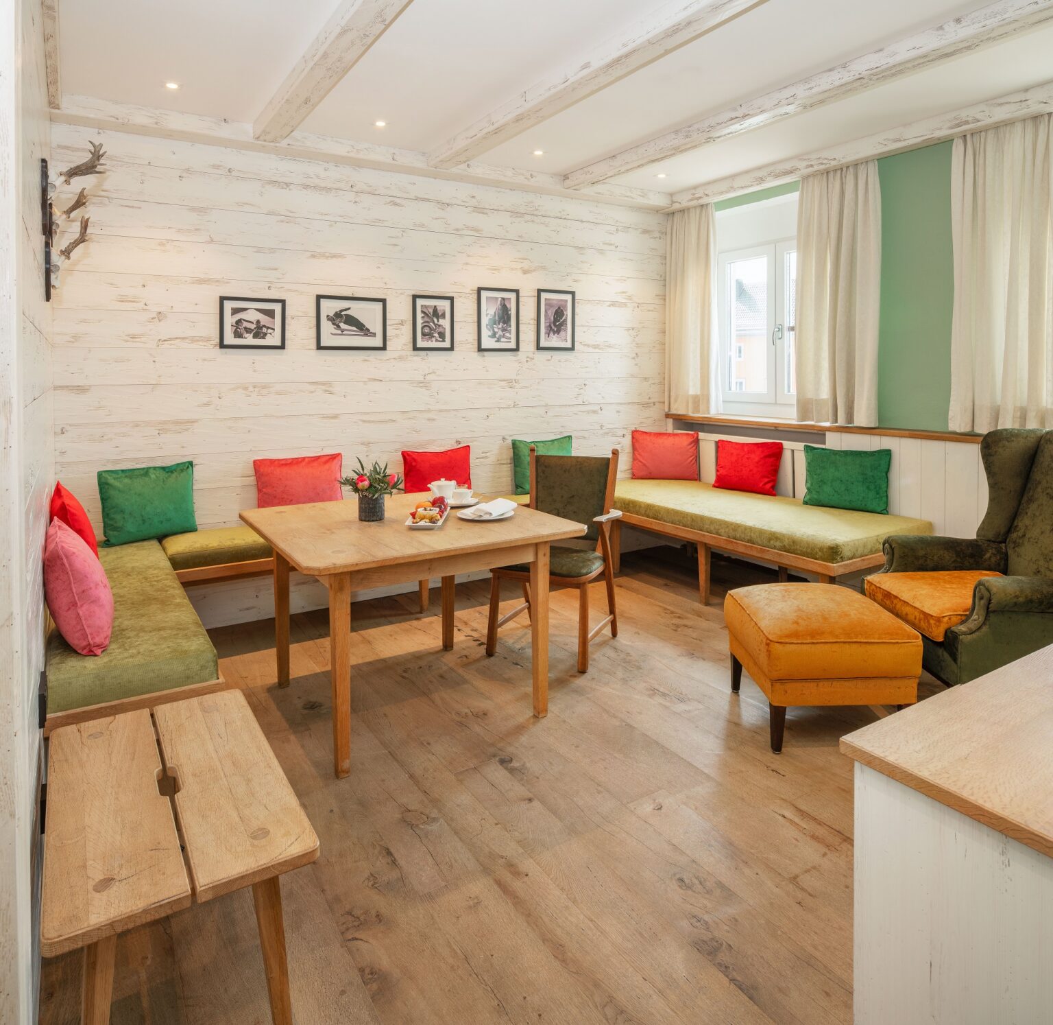 Wohnzimmer in der Juniorsuite mit Eckbank mit vielen bunten Kissen, Tisch, Sessel mit Fußhocker, Parkett, weiße Holzwände, Schwarzweiß-Fotos und Geweihe an den Wänden