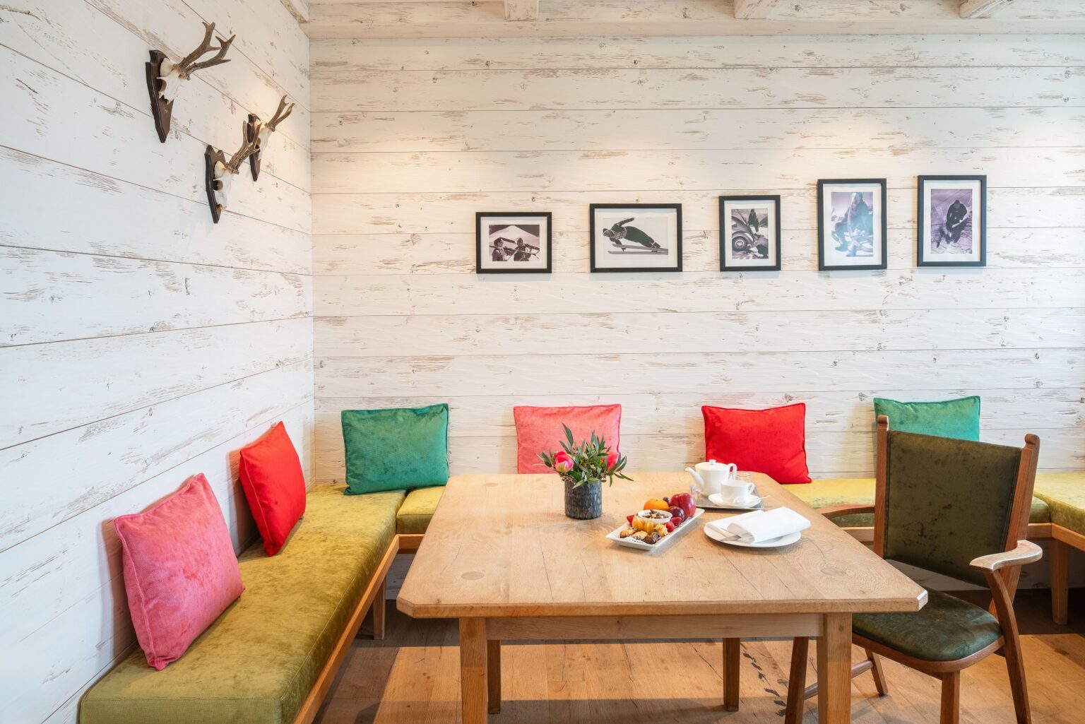 Wohnzimmer in der Juniorsuite mit Eckbank mit vielen bunten Kissen, Tisch, Sessel mit Fußhocker, Parkett, weiße Holzwände, Schwarzweiß-Fotos und Geweihe an den Wänden
