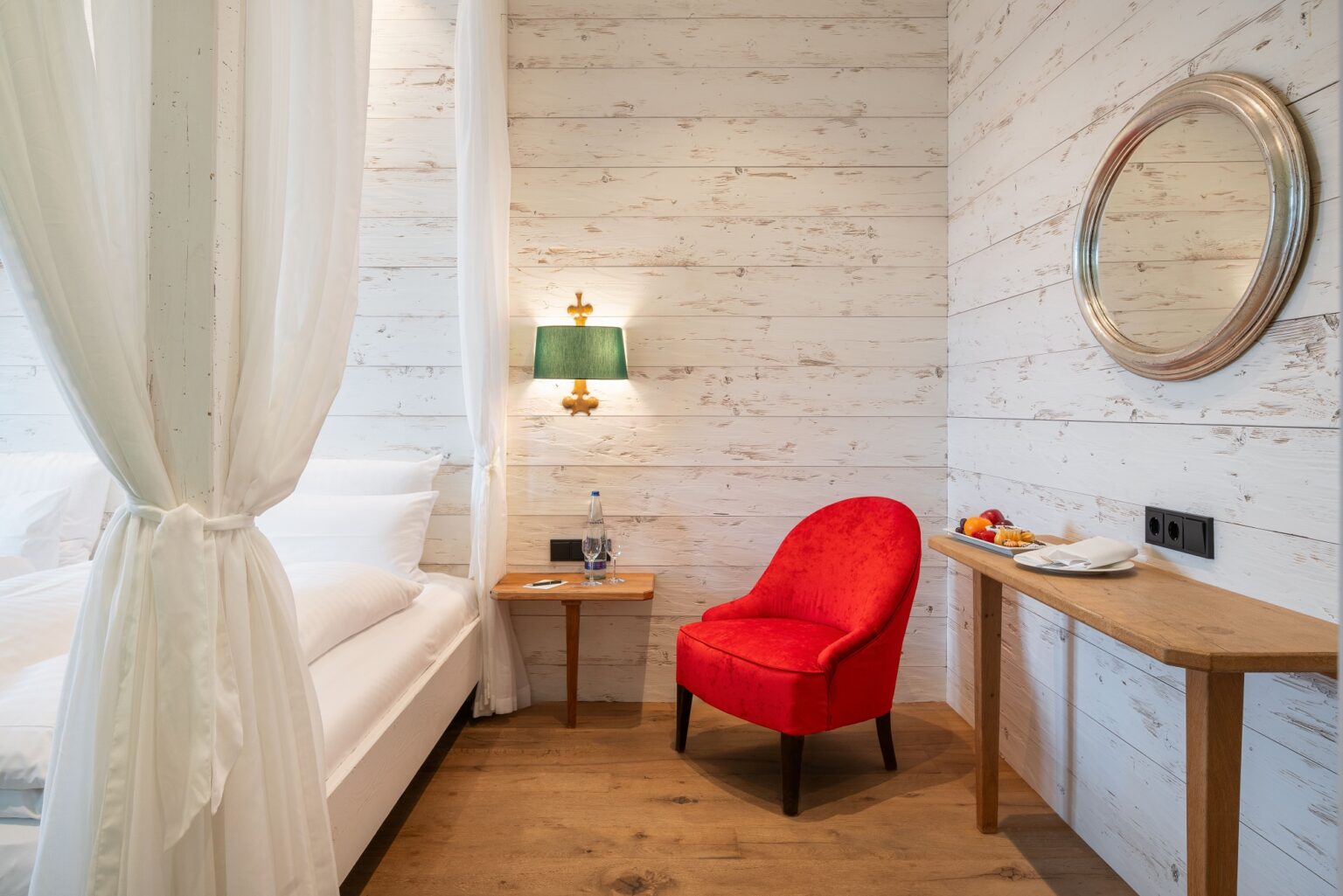 Schlafzimmer Juniorsuite, Himmelbett mit weißem Vorhang, roter Sessel, Spiegel und Konsolentisch rechts, Wandbeleuchtung mit grünem Schirm, weiße Holzwände, Parkett